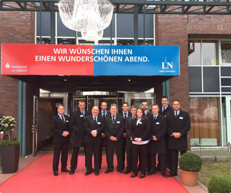 Eine Gruppe von Sicherheitskräften in dunklen Anzügen steht auf einem roten Teppich vor einem Gebäudeeingang mit einem Willkommensbanner.