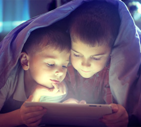 Kinder mit Tablet unter Decke Zwei Kinder liegen unter einer Decke und blicken konzentriert auf ein leuchtendes Tablet in einer dunklen Umgebung.