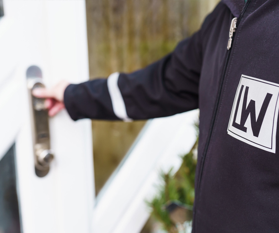 Eine Person in dunkler Jacke mit dem Logo LW greift nach dem Türgriff einer weißen Haustür.