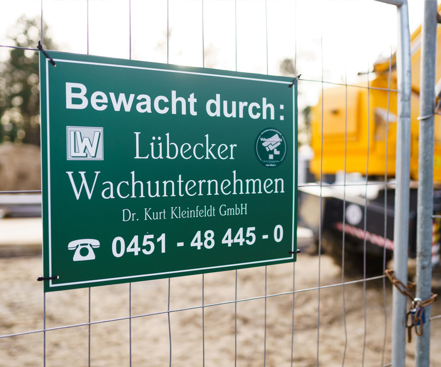 Grünes Hinweisschild mit der Aufschrift „Bewacht durch: Lübecker Wachunternehmen“ an einem Drahtzaun vor einer Baustelle mit gelbem Baufahrzeug.