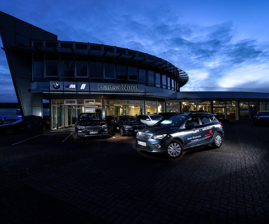 Beleuchtetes Autohaus bei Dämmerung mit mehreren Fahrzeugen, darunter ein Wagen mit der Aufschrift Lübecker Wachunternehmen.