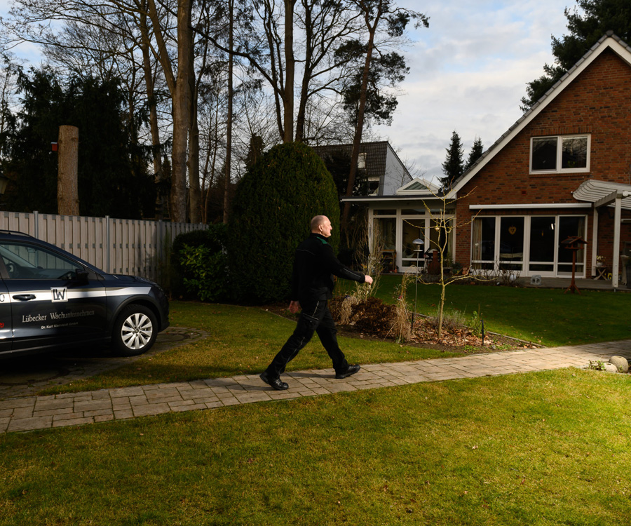 Ein Sicherheitsmitarbeiter in dunkler Uniform geht auf einem gepflasterten Weg an einem Wohnhaus vorbei, neben ihm parkt ein dunkles Einsatzfahrzeug.