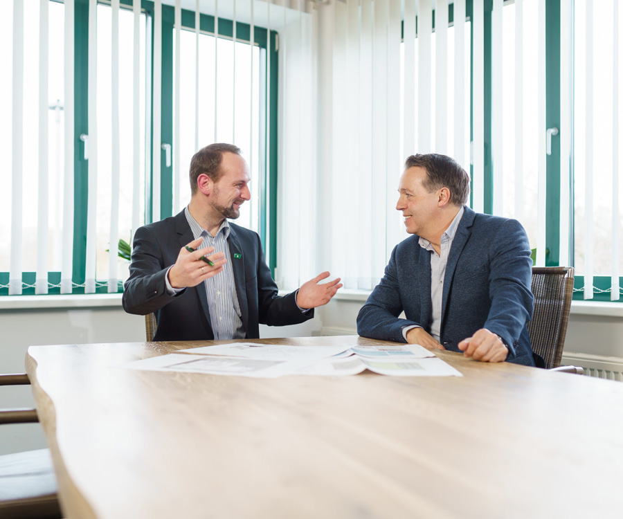 Zwei Männer in Business-Kleidung sitzen an einem Holztisch und besprechen Dokumente in einem hellen Büro mit großen Fenstern.