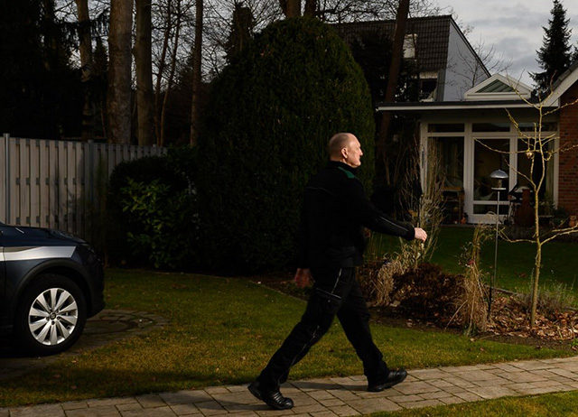 Ein Sicherheitsmitarbeiter in dunkler Uniform geht auf einem gepflasterten Weg an einem Wohnhaus und einem beschrifteten Einsatzfahrzeug vorbei.