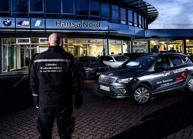 Ein Sicherheitsmitarbeiter in dunkler Uniform blickt bei Dämmerung auf ein beleuchtetes Autohaus und ein davor geparktes Einsatzfahrzeug.