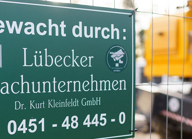 Grünes Hinweisschild mit der Aufschrift „Bewacht durch: Lübecker Wachunternehmen“ an einem Bauzaun vor einem gelben Baufahrzeug.