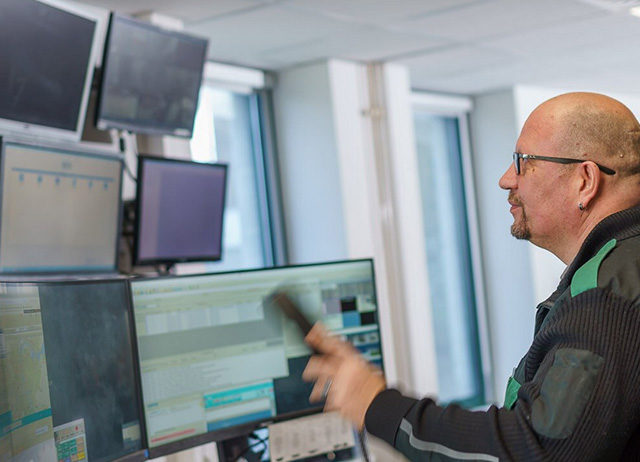 Ein Mann mit Brille in dunkler Arbeitskleidung blickt in einer Leitstelle auf mehrere Computerbildschirme mit verschiedenen Datenanzeigen.