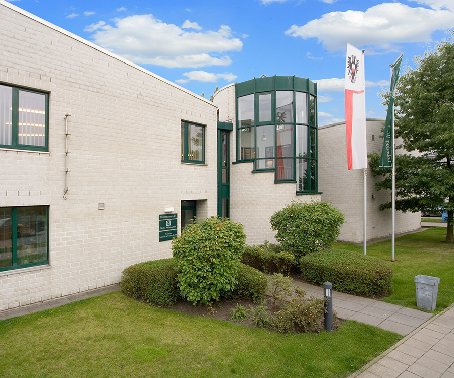 Modernes Bürogebäude aus hellem Backstein mit grünen Fensterrahmen, einem runden Glasvorbau und gepflegten Grünanlagen unter blauem Himmel.