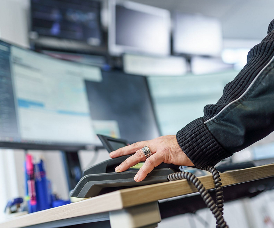 Eine Hand mit einem Ring ruht auf einem Telefonhörer vor mehreren Computerbildschirmen in einer Leitstelle.
