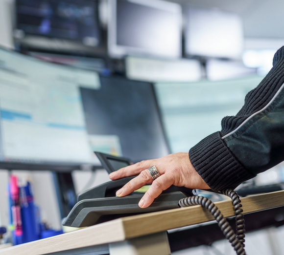 Eine Hand mit einem gemusterten Ring ruht auf einem schwarzen Telefonhörer vor mehreren unscharfen Computerbildschirmen.
