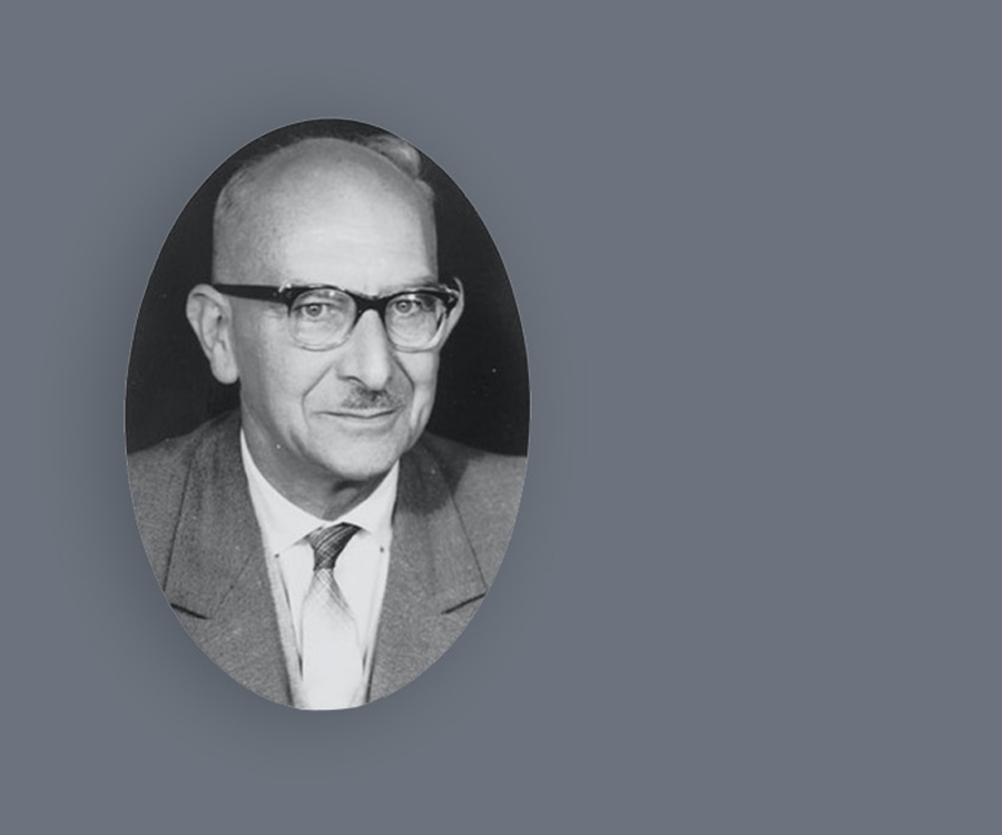 Ein älterer Mann mit Brille und Schnurrbart in einem Anzug, dargestellt in einem ovalen Porträtausschnitt auf grauem Hintergrund.