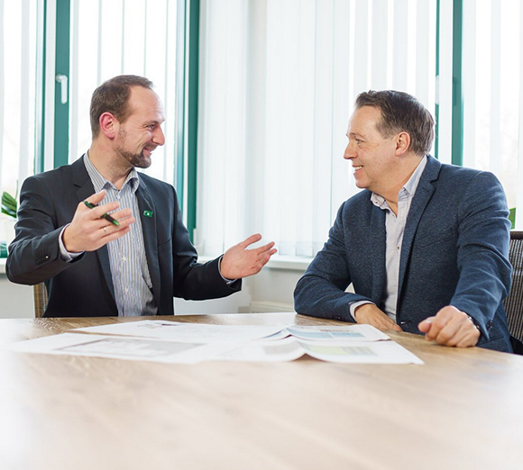 Zwei Männer in Business-Kleidung sitzen an einem Holztisch und führen ein Gespräch über ausgelegte Dokumente.
