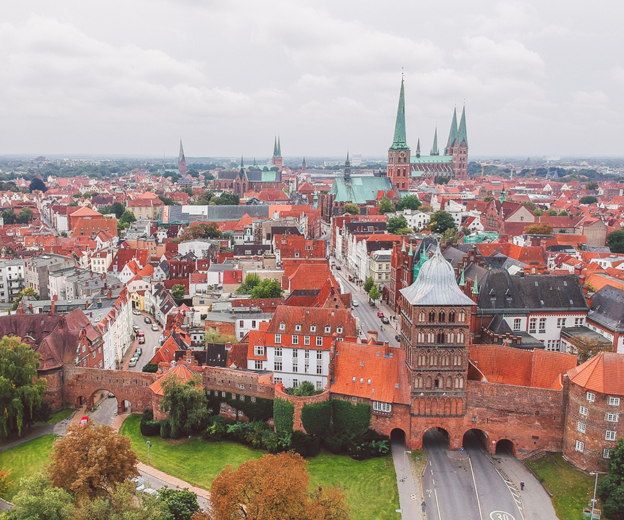 Vogelperspektive auf eine europäische Altstadt mit roten Ziegeldächern, einem historischen Stadttor aus Backstein und hohen Kirchtürmen in der Ferne.