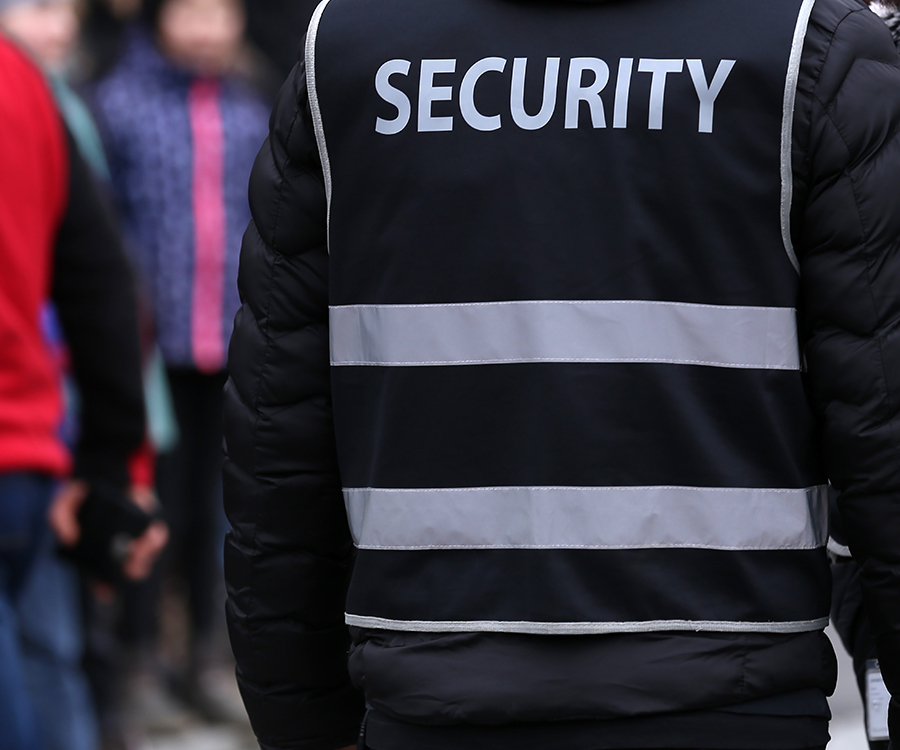 Rückansicht einer Person in einer schwarzen Weste mit der Aufschrift SECURITY und zwei horizontalen Reflexstreifen vor unscharfem Hintergrund.