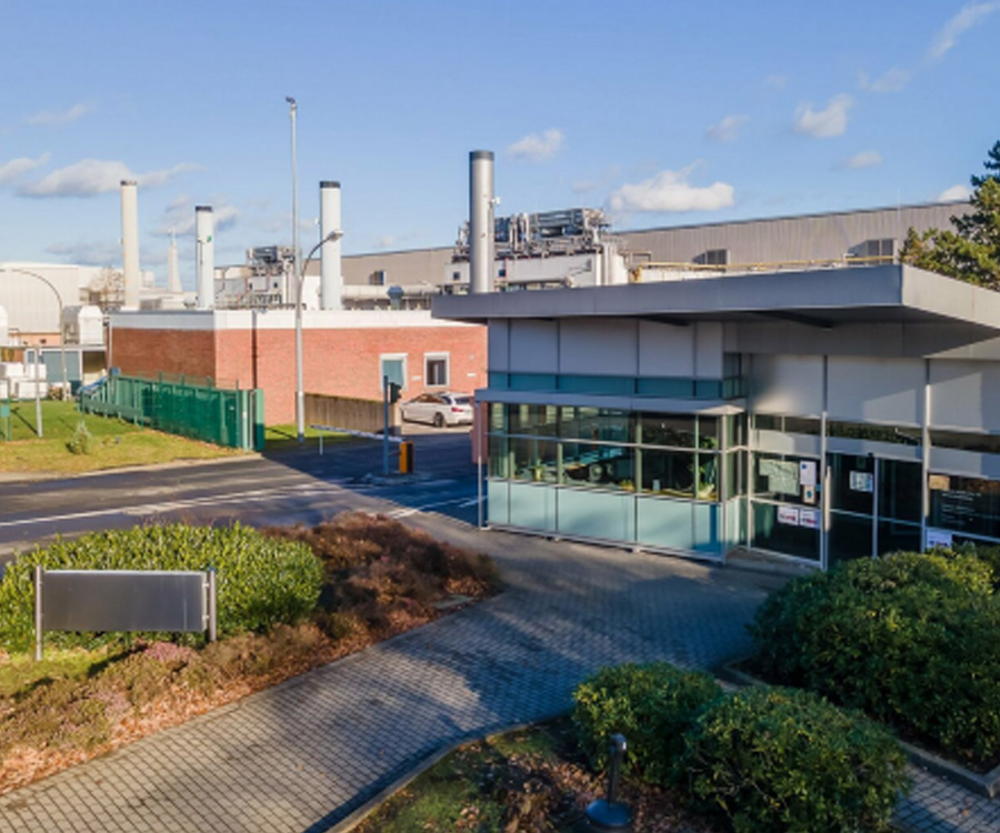 Ein modernes Pförtnergebäude an der Einfahrt zu einem weitläufigen Industriegelände mit Fabrikhallen und Schornsteinen im Hintergrund.