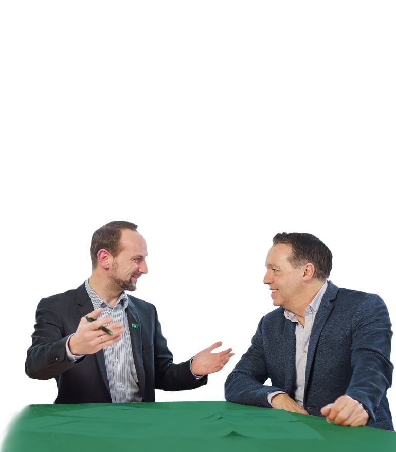 Zwei Männer in Sakkos sitzen an einem grünen Tisch und führen ein Gespräch, wobei einer mit einem Stift gestikuliert.