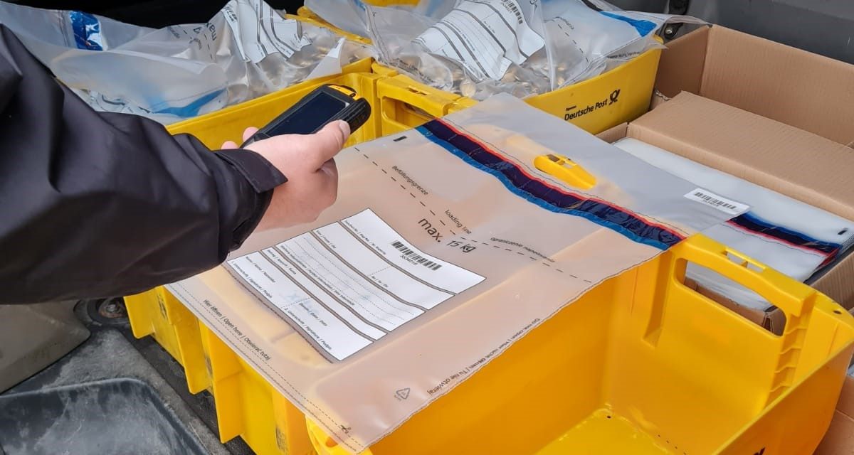 Eine Hand hält ein Scangerät über gelbe Kunststoffkisten mit gefüllten Sicherheitsbeuteln im Laderaum eines Transporters.