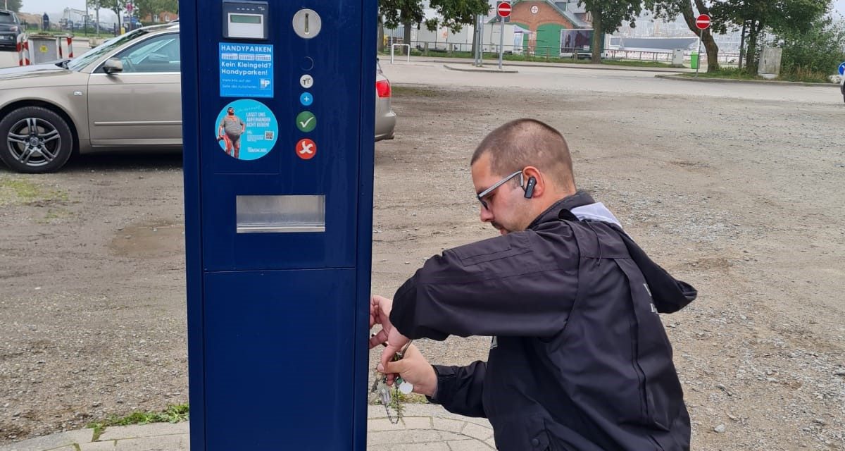 Ein Mann in dunkler Arbeitskleidung kniet neben einem blauen Parkscheinautomaten und öffnet diesen mit einem Schlüsselbund.