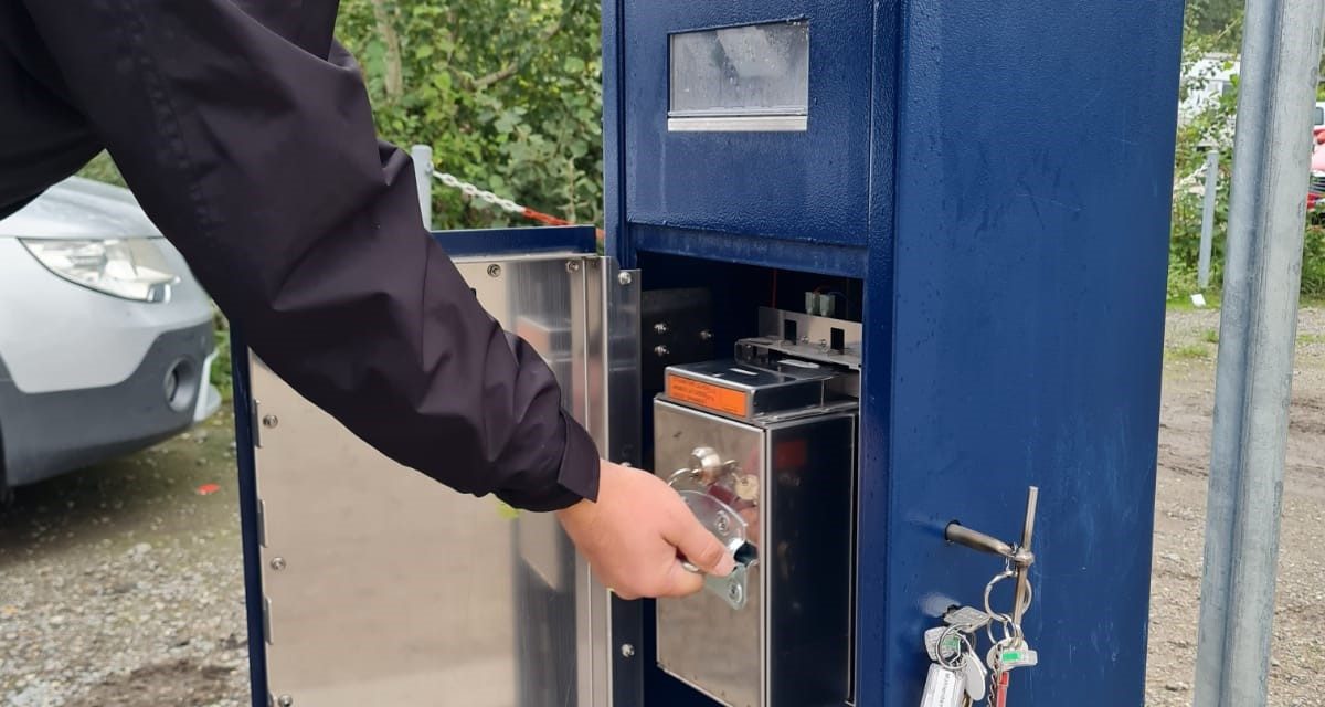 Ein Mann öffnet die Wartungsklappe eines blauen Parkscheinautomaten und bedient einen Verschluss an der internen Metallbox.