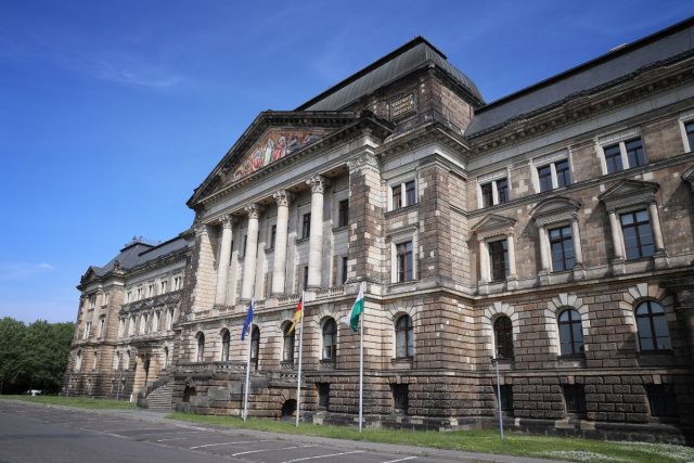 Großes historisches Steingebäude mit Säulenportikus, Giebelrelief und mehreren Flaggen vor blauem Himmel.