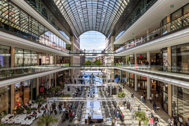 Innenansicht eines mehrstöckigen Einkaufszentrums mit einem großen Glasdach, Galerien und einem Veranstaltungsbereich im Erdgeschoss.