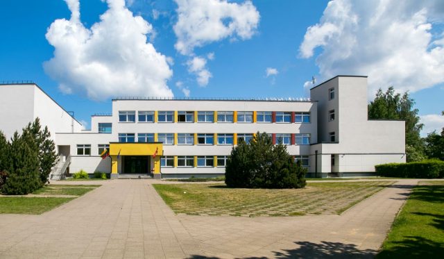 Modernes, mehrstöckiges Schulgebäude mit weißer Fassade, farbigen Fensterumrandungen und einem gepflasterten Vorplatz unter blauem Himmel.