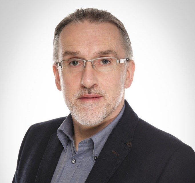 Ein Mann mit Brille, kurzem grauem Haar und Bart trägt ein blaues Hemd sowie ein dunkles Sakko vor hellem Hintergrund.