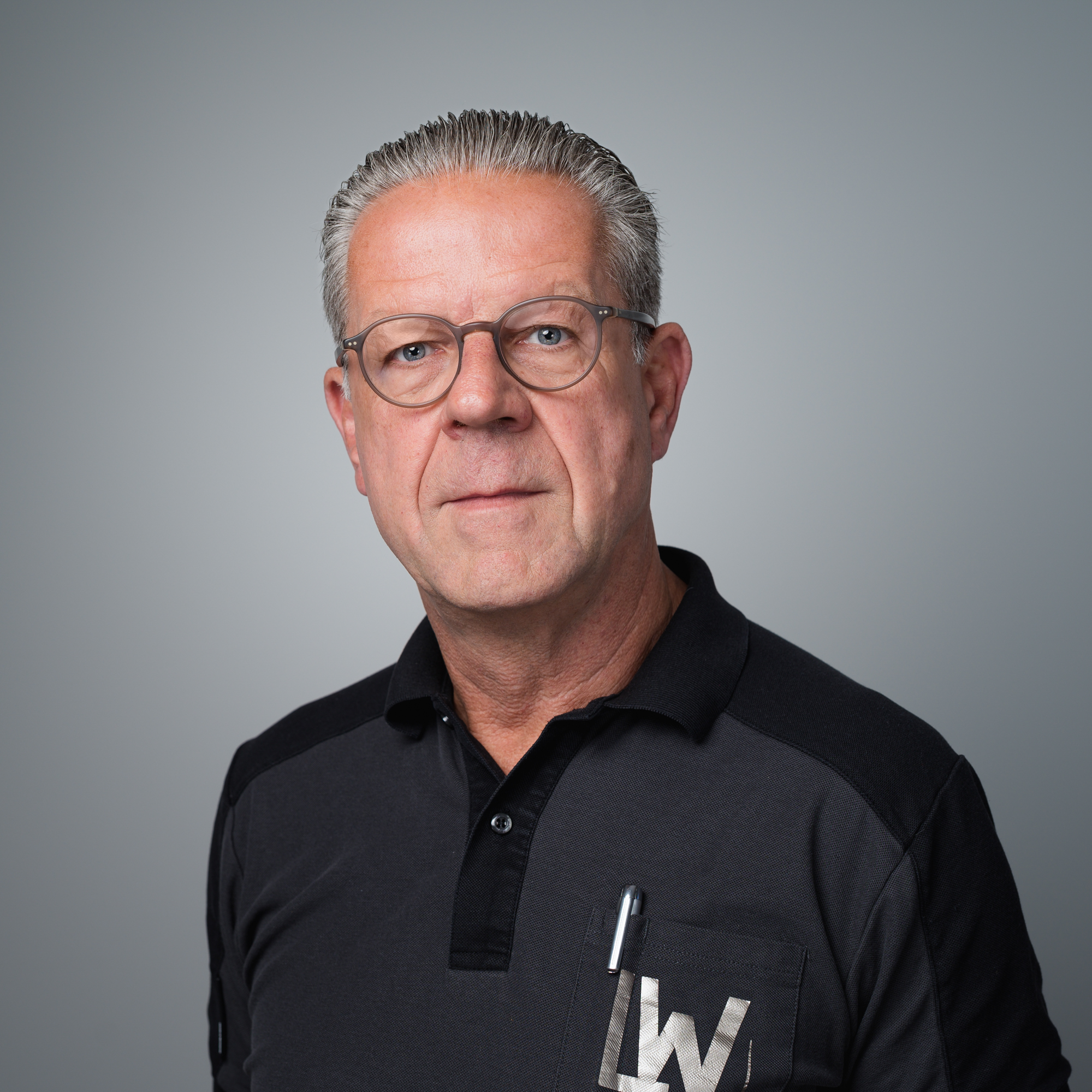 Porträt eines Mannes mit Brille und kurzem grauen Haar in einem dunklen Poloshirt vor grauem Hintergrund.
