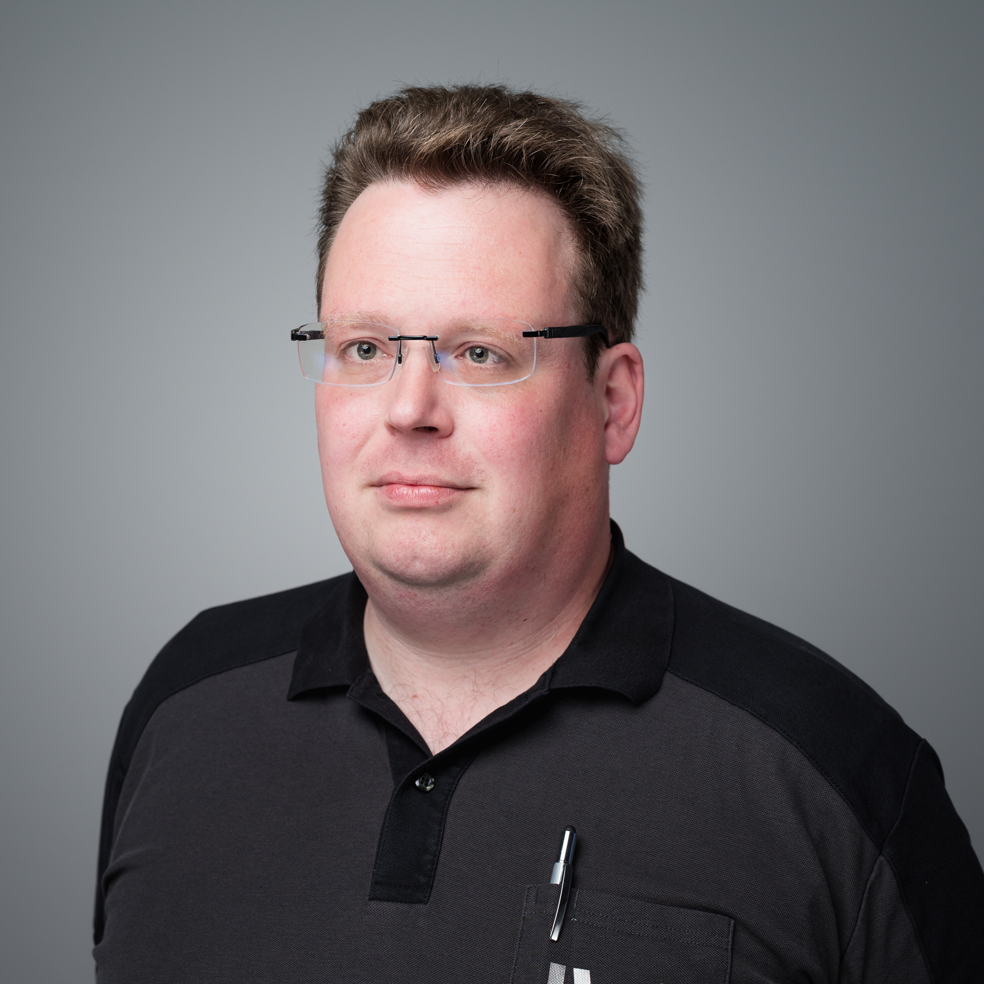 Ein Mann mit Brille und kurzem braunem Haar trägt ein dunkles Poloshirt vor einem grauen Hintergrund.