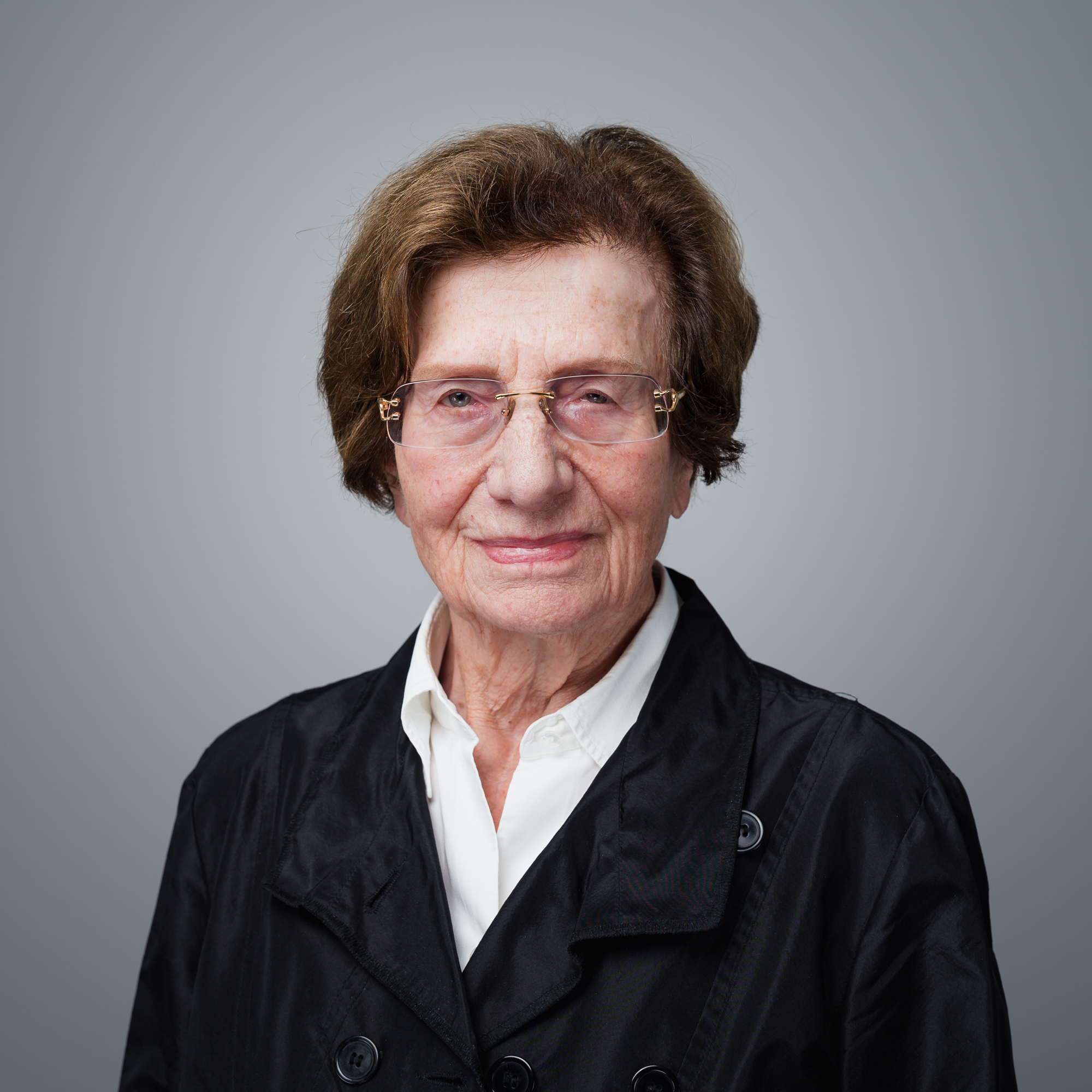 Eine ältere Frau mit kurzen braunen Haaren und Brille trägt eine weiße Bluse sowie eine schwarze Jacke vor einem grauen Hintergrund.