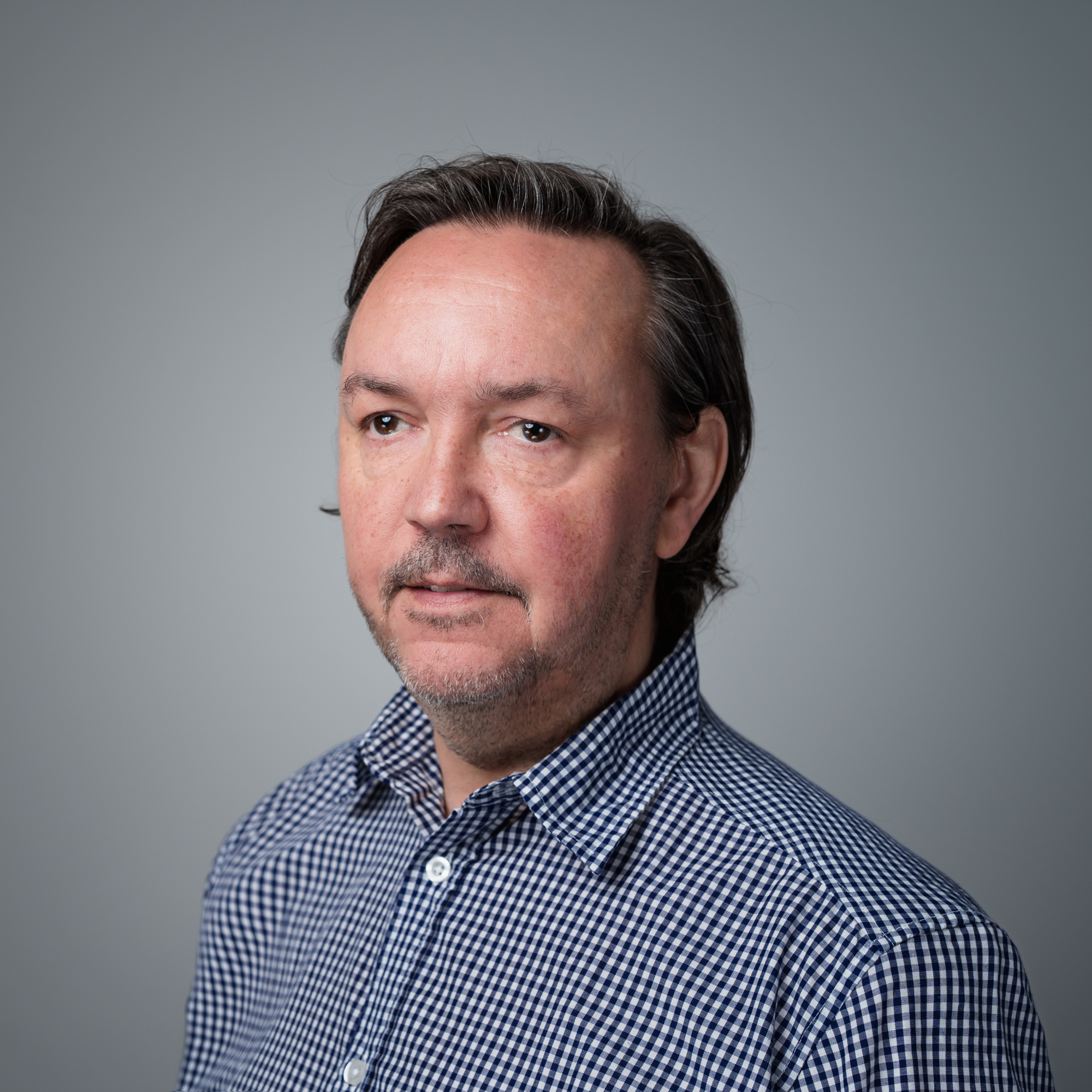 Ein Mann mit dunklen Haaren und kurzem Bart trägt ein blau-weiß kariertes Hemd vor einem grauen Hintergrund.
