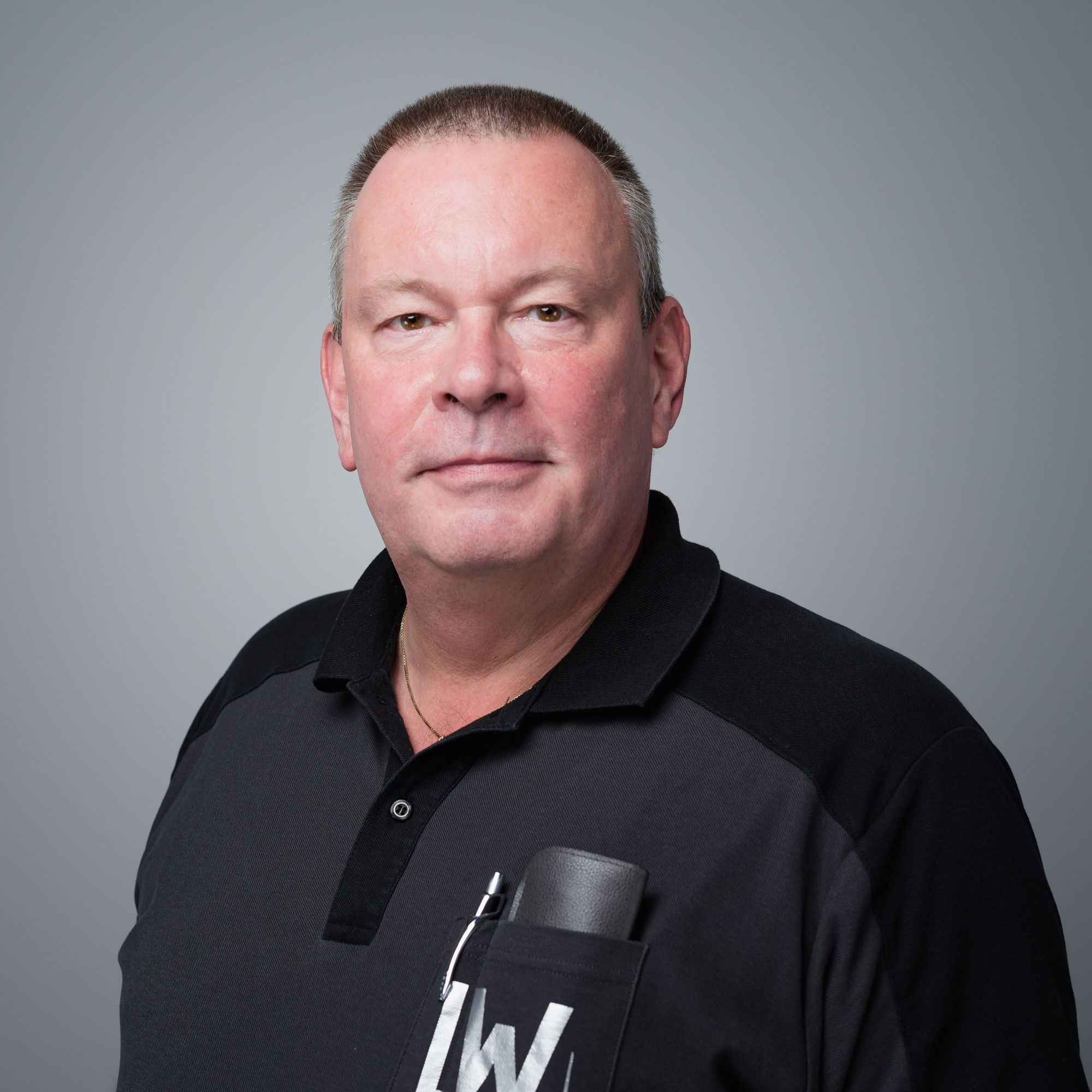 Ein Mann mit kurzen Haaren in einem dunklen Poloshirt vor einem neutralen grauen Hintergrund.