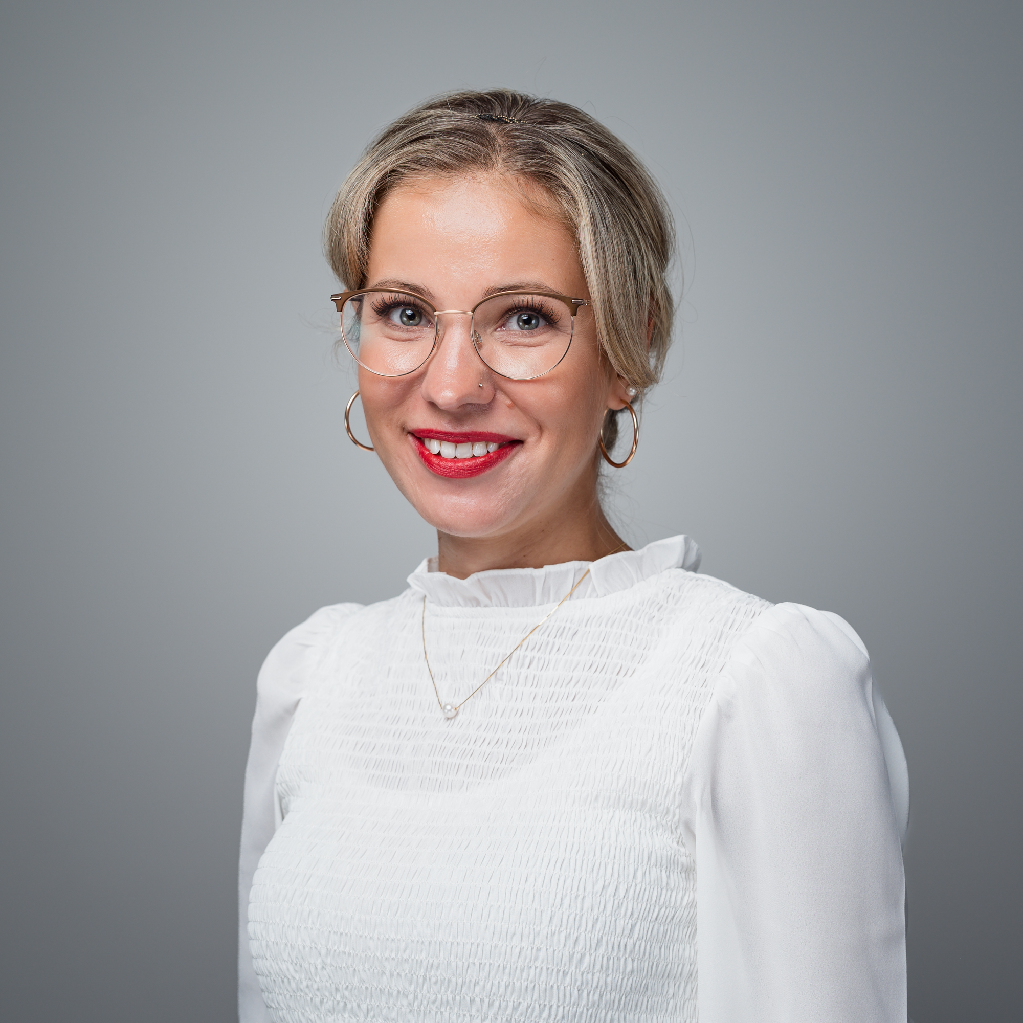 Eine lächelnde Frau mit Brille, blondem Haar und rotem Lippenstift trägt eine weiße Bluse vor einem grauen Hintergrund.