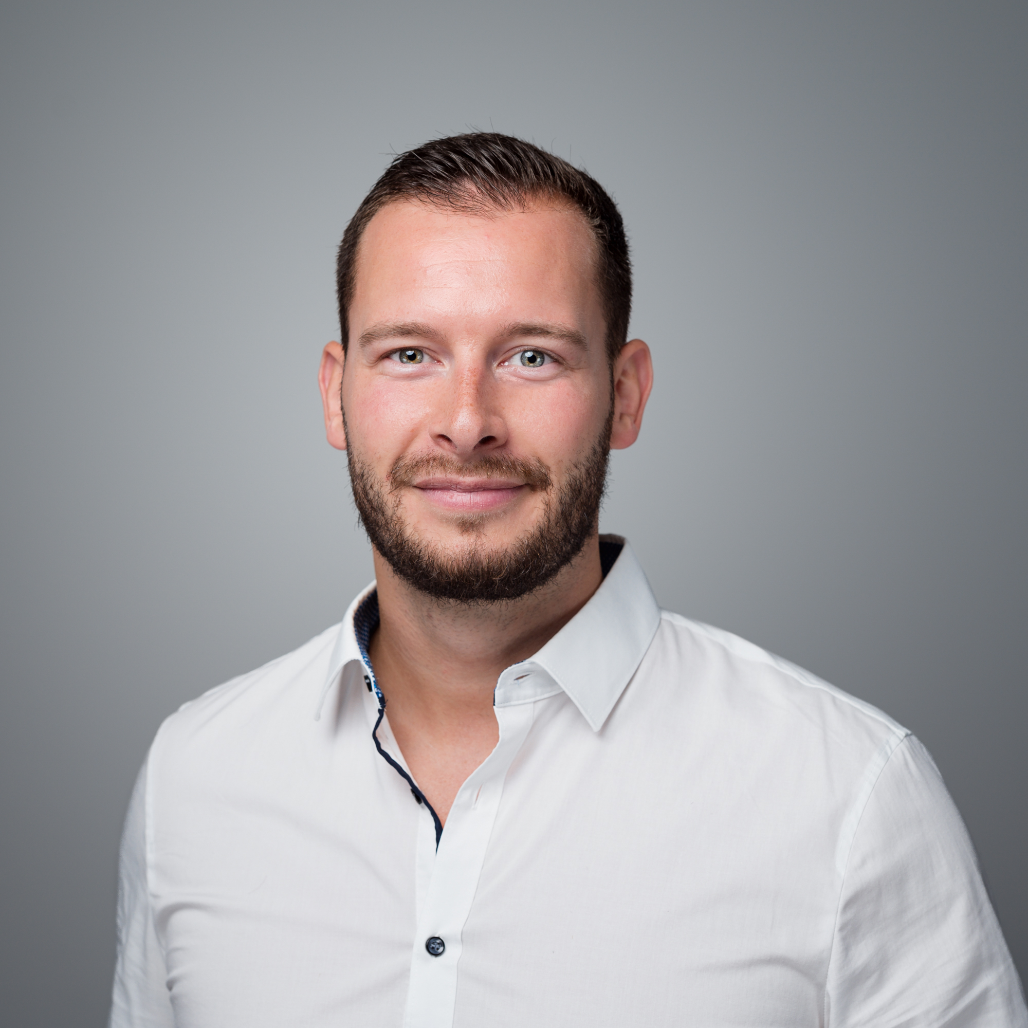 Ein lächelnder Mann mit kurzem braunem Haar und Bart trägt ein weißes Hemd vor einem grauen Hintergrund.