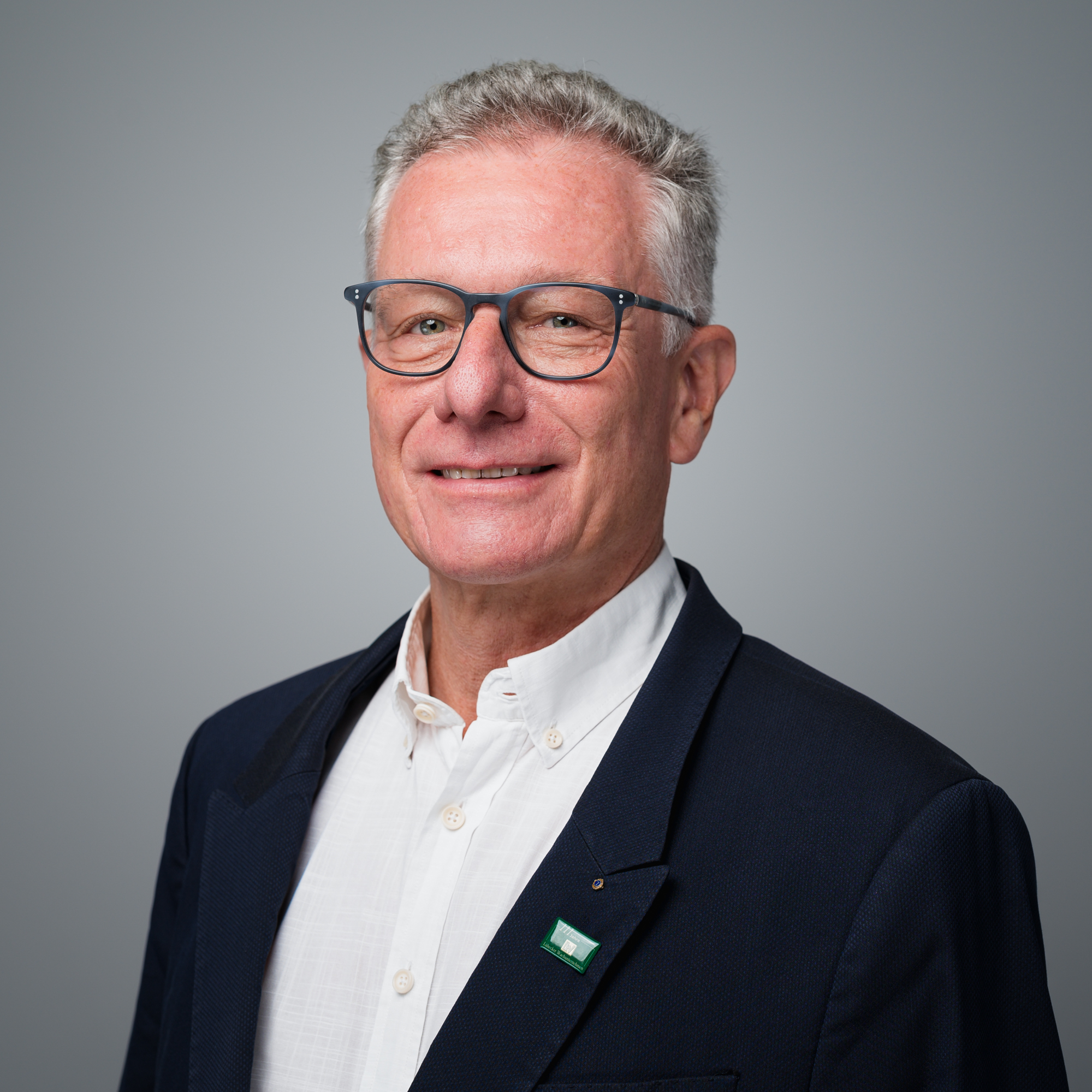 Ein lächelnder Mann mit grauen Haaren und Brille trägt ein weißes Hemd und ein dunkelblaues Sakko vor einem neutralen grauen Hintergrund.