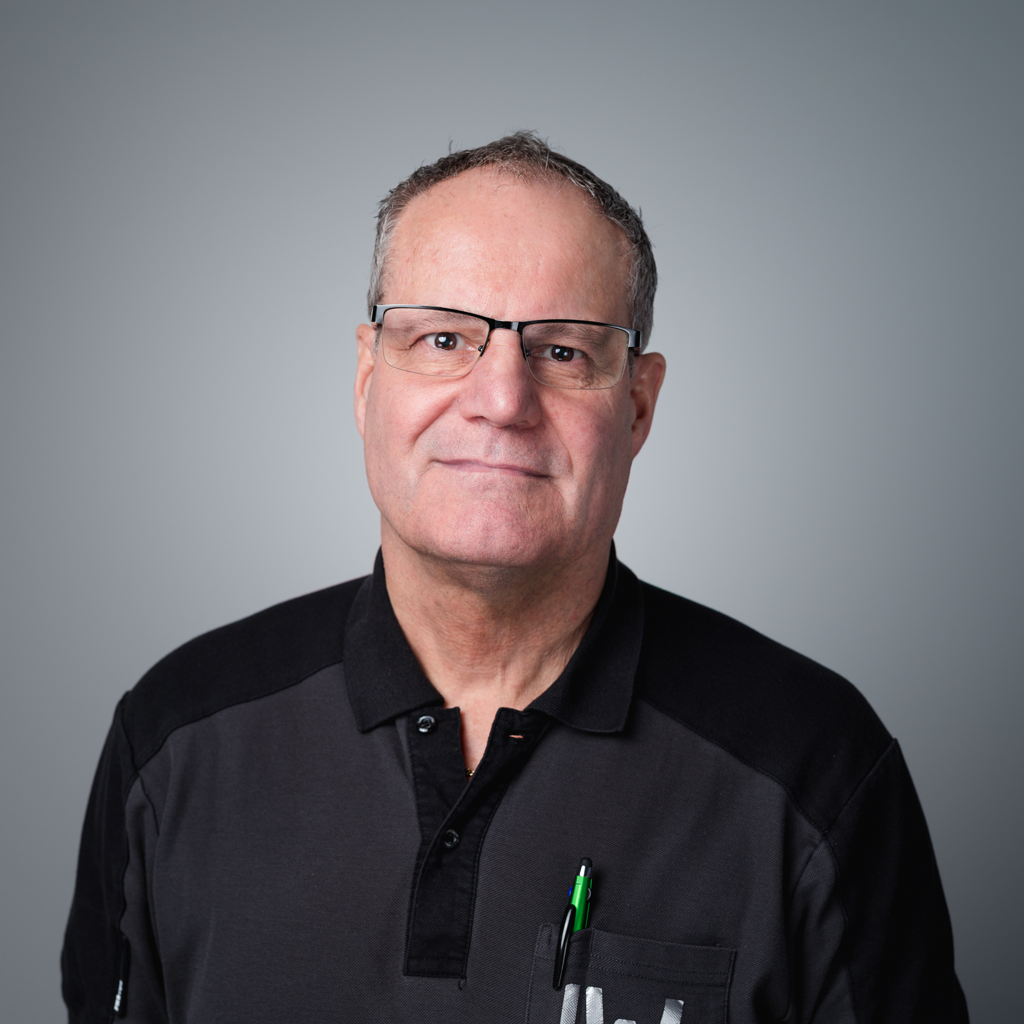 Ein Mann mit Brille und kurzem grauen Haar trägt ein dunkles Poloshirt vor einem neutralen grauen Hintergrund.