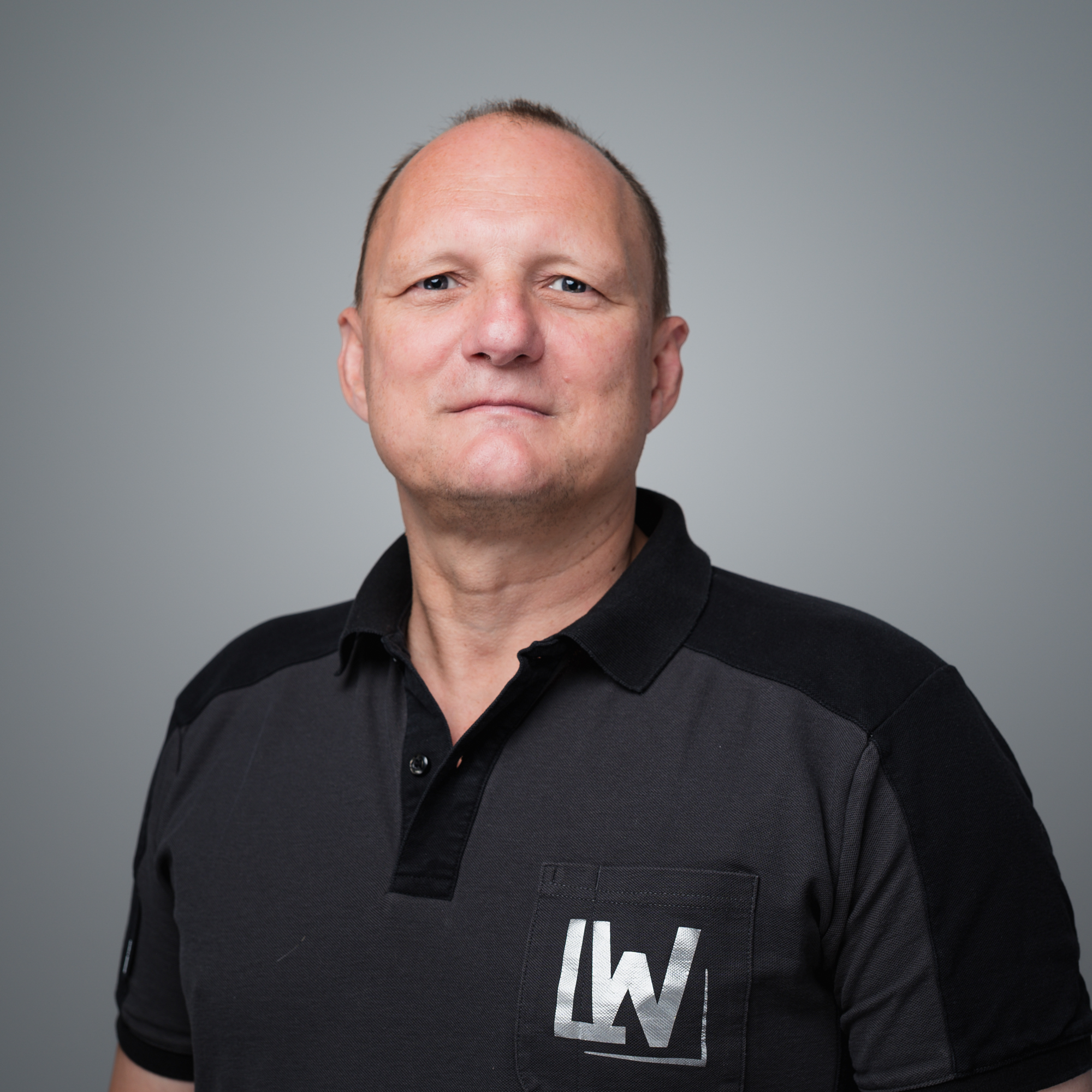 Ein Mann mit kurzem Haar trägt ein dunkles Poloshirt mit dem Aufdruck LW vor einem grauen Hintergrund.