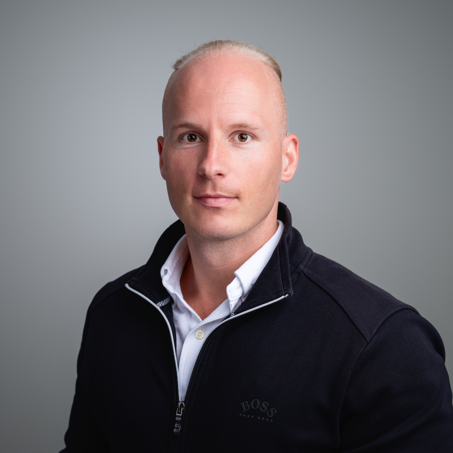 Ein Mann mit kurzen blonden Haaren trägt ein weißes Hemd und eine dunkle Jacke vor einem grauen Hintergrund.