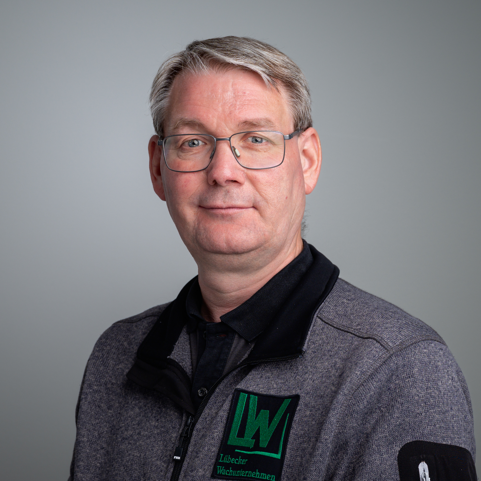 Ein Mann mit Brille und kurzem grauem Haar trägt eine graue Fleecejacke mit dem Logo LW Lübecker Wachunternehmen.