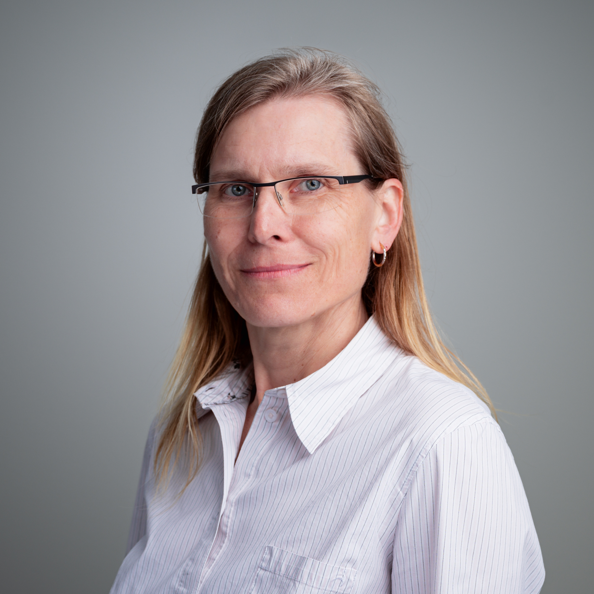 Eine Frau mit Brille und hellen Haaren trägt eine weiß gestreifte Bluse vor einem grauen Hintergrund.