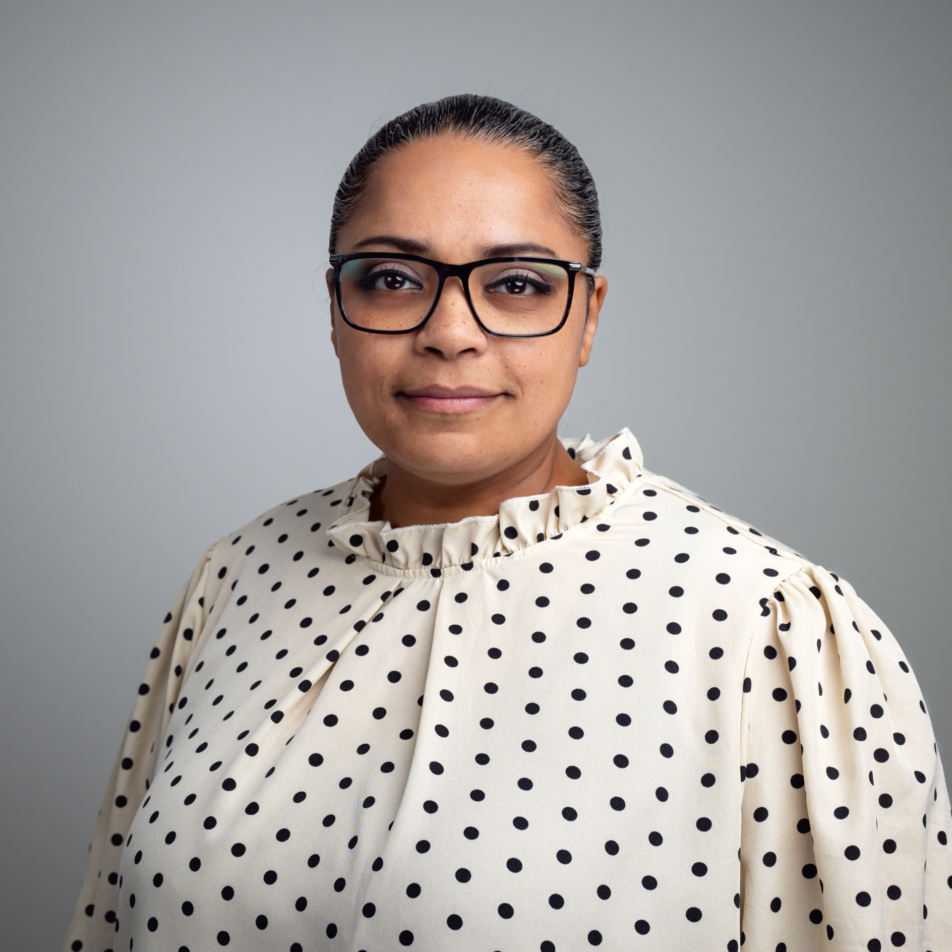 Eine Frau mit Brille und zurückgebundenem Haar trägt eine helle Bluse mit schwarzen Punkten vor einem grauen Hintergrund.