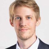 Ein blondhaariger Mann mit blauen Augen trägt ein hellblaues Hemd und ein dunkles Sakko vor einem hellen Hintergrund.