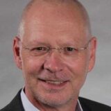 Ein lächelnder Mann mit Glatze und randloser Brille trägt ein dunkles Sakko vor einem grauen Hintergrund.