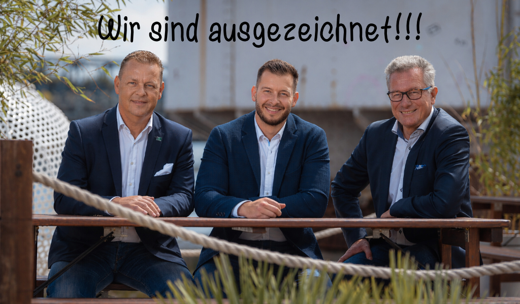 Drei lächelnde Männer in dunklen Sakkos sitzen nebeneinander an einem Holztisch im Freien.